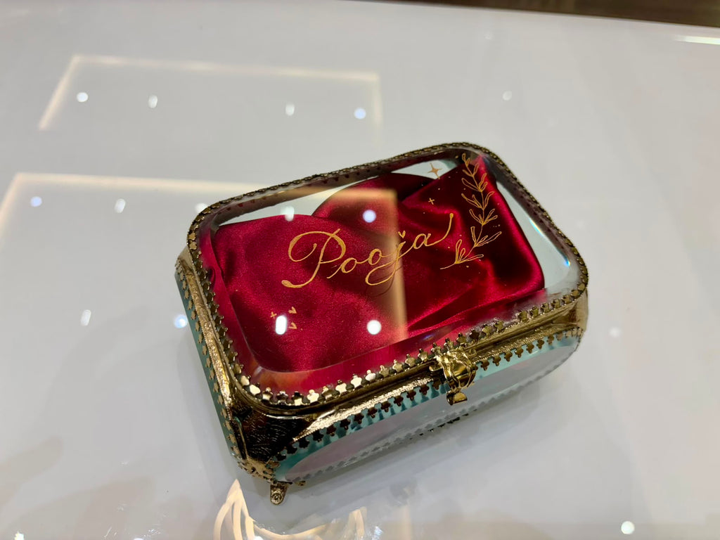 A rectangular glass trinket box with ornate gold-tone metal trim and a mirror interior, featuring the name 'Pooja' in gold cursive script on the lid.